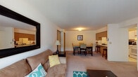 Anderson Apartments presents a thoughtfully laid-out single-floor residence designed for comfortable everyday living. The open-concept living and dining area forms the heart of the unit, where plush carpeting and neutral walls create a warm, inviting atmosphere. The living room offers generous space for seating and entertainment, highlighted by a sliding glass door with vertical blinds that fills the room with natural light. Adjacent to the living area, the dining space accommodates a full-sized table and chairs, seamlessly connecting to a breakfast bar that bridges the kitchen. The kitchen is efficiently designed with white cabinetry trimmed in warm wood tones, ample counter space, and tile flooring, providing a clean and functional cooking environment. Two well-proportioned bedrooms each feature plush carpet, natural window light, and closet storage. The primary bedroom includes dual dark-wood dressers with mirrors, lending an elegant, cohesive look. A secondary room serves as a flexible office or den space, complete with a closet and window. The bathroom is straightforward and functional, featuring tile flooring, a vanity with storage, and a shower-tub combination. Throughout the residence, a consistent neutral color palette and carpet flooring contribute to a cohesive, comfortable feel.