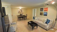 This single-floor space presents an open-concept layout that seamlessly integrates the living and dining areas into one cohesive environment. The living area is anchored by a plush, light-colored sofa dressed with decorative pillows in neutral and blue tones, complemented by a dark-stained wooden coffee table with a tray-style top that adds a sophisticated touch. A patterned accent chair rounds out the seating arrangement, contributing to an inviting and functional layout.

The dining area features a dark-finished round table surrounded by matching cross-back chairs, creating an elegant and intimate space for meals. A framed botanical artwork above a chair rail adds a classic decorative element to the wall. On the opposite side, a curated gallery of four vibrant abstract artworks introduces a pop of color and visual interest to the warm, beige-toned walls.

Soft, neutral carpeting flows throughout the space, enhancing the warm ambiance. Crown molding runs along the ceiling perimeter, lending a refined architectural detail. A window in the background allows natural light to filter in, brightening the interior. The overall atmosphere is calm, classic, and well-appointed.