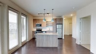 This 2-bedroom, 2-bathroom residence offers a thoughtfully designed single-level layout that blends functionality with modern style. The open-concept kitchen and living area serve as the heart of the home, anchored by a large central island topped with granite countertops and illuminated by a pair of pendant lights in a muted teal finish. The kitchen is well-appointed with wood-toned cabinetry, stainless steel appliances including a refrigerator, stove, and microwave, and a clean tile backsplash that adds a crisp contrast to the warm cabinetry tones. Dark hardwood flooring extends throughout the main living area, providing a rich, grounded aesthetic that complements the light, neutral wall colors. A sliding glass door allows natural light to pour into the space, enhancing the open and airy atmosphere. The layout flows seamlessly from the kitchen into the living area and beyond toward the two bedrooms, offering a practical and comfortable arrangement. The overall design reflects a contemporary aesthetic with warm wood accents, quality finishes, and an efficient use of space that caters to everyday living.