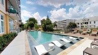 This outdoor amenity space centers around a striking resort-style swimming pool designed for both relaxation and visual appeal. The pool features clean, geometric lines with crystal-clear turquoise water, framed by a smooth, light-toned deck that extends generously around the perimeter. A set of sleek, sculptural white chaise loungers lines the poolside, offering a modern aesthetic alongside practical comfort for sunbathing and leisure. Accompanying small round side tables provide convenient surfaces for poolside use. The surrounding landscape is lush with mature trees, lending a sense of natural privacy and shade. A railed perimeter defines the space while maintaining open sightlines to the verdant surroundings. Additional seating and lounging areas are positioned throughout the deck, including a raised platform area that offers alternative vantage points. The open sky above allows abundant natural light to flood the space, creating a bright and inviting atmosphere throughout the day. The overall design balances contemporary architecture with a welcoming, resort-inspired ambiance, making it a well-appointed outdoor retreat well-suited for both active use and quiet relaxation.