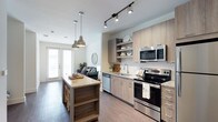 This single-floor, one-bedroom, one-bathroom residence offers a thoughtfully designed open-concept layout that seamlessly connects the kitchen and living areas. The kitchen is a standout feature, showcasing light wood cabinetry, open shelving, and a granite-topped island with decorative spindle detailing and built-in storage below. Pendant lights hang above the island, complementing the track lighting overhead to create a warm, layered lighting scheme. Stainless steel appliances, including a range, dishwasher, and over-the-range microwave, provide a modern, functional aesthetic, while a subway tile backsplash adds a clean, classic touch. Durable wood-look flooring runs throughout, adding warmth and continuity across the space. The open flow into the living area allows natural light to pour in through glass-paneled doors at the far end, creating a bright and airy ambiance. The overall design balances contemporary style with practical functionality, making this residence a comfortable and visually cohesive living space.