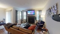 Located at 9806 Mahogany Drive in Gaithersburg, MD, this single-floor residence offers a well-organized layout with distinct living, dining, kitchen, bedroom, and bathroom areas. The living room serves as the heart of the home, anchored by a classic fireplace and flanked by multiple windows that invite natural light throughout the day. The space accommodates comfortable seating and wall-mounted media, creating a functional yet welcoming atmosphere with hardwood-style flooring underfoot. Adjacent to the living room, the dining area features a sliding glass door that opens to a view of lush greenery outside, providing a bright, airy setting ideal for meals and gatherings. An overhead drum pendant light adds a modern touch to the space. The kitchen is efficiently designed with warm honey-toned cabinetry, white countertops, a tile backsplash, and black appliances including a range, dishwasher, and refrigerator. A pass-through window connects the kitchen to the adjacent area, enhancing the open feel. The bedroom is a calm retreat finished with soft grey walls, plush carpet, and a sliding glass door offering direct access to outdoor greenery. The full bathroom is clean and functional, featuring a single-sink vanity with white countertop, a large mirror, and a shower-tub combo.