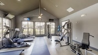Located at 230 City Green Way in Chattanooga, Tennessee, this well-appointed fitness center offers a comprehensive workout facility designed to accommodate a wide range of exercise needs. The main exercise area is defined by soaring vaulted ceilings, which lend an open, airy quality to the space, while ceiling fans help maintain a comfortable environment during workouts. Large, multi-pane windows line the walls, flooding the interior with abundant natural light and providing views of the surrounding greenery outdoors.

The cardio zone is generously equipped with multiple treadmills and elliptical trainers arranged in rows facing the windows, allowing users to exercise while enjoying the outdoor scenery. Wall-mounted televisions are positioned throughout for entertainment during workouts. The strength training section features a selection of cable and resistance machines arranged across durable, low-pile fitness flooring, providing ample space for both machine-based and free-weight exercises.

A dedicated kitchenette area complements the fitness space, outfitted with dark wood cabinetry, a stainless steel sink, countertop workspace, and a compact refrigerator — ideal for storing refreshments. The neutral gray color palette throughout the facility gives the space a clean, modern aesthetic. Recessed lighting and a clearly marked exit contribute to a safe and functional environment for all users.
