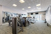 Located at 170 Market St in Manassas Park, VA, this well-appointed fitness center offers a generous, open floor plan thoughtfully designed to accommodate a wide range of exercise needs. The space is finished with durable, patterned carpet flooring throughout and a dropped ceiling with recessed panel lighting that ensures consistent, bright illumination across the entire area.

The cardio section features a row of TRUE-brand treadmills and rowing machines, positioned along a full-length wall mirror that enhances the sense of space. Strength training is comprehensively covered with a selection of TRUE selectorized cable and resistance machines, a power rack with free weights and barbells, adjustable benches, and a dumbbell rack. A dedicated functional training zone includes stability balls, medicine balls, resistance bands, and additional benches for versatile workout options.

A large, dramatic abstract wall mural serves as a striking focal point, adding an energetic and artistic ambiance to the environment. Large windows near the entrance allow natural light to filter in, offering views of the surrounding area. Additional amenities include a water fountain with a bottle-filling station. The facility is designed to support a full-body workout experience in a clean, modern, and motivating setting.