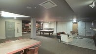 This single-floor recreational lounge is thoughtfully designed to accommodate a variety of social and leisure activities. The space is anchored by a full-size pool table positioned at its center, illuminated by a stylish pendant light that draws attention to the focal point of the room. The dark carpet flooring and grey-toned ceiling create a warm, intimate atmosphere throughout.

To one side, a casual lounge area is arranged with contemporary seating, including armchairs and accent chairs set atop a patterned area rug, offering a comfortable spot for conversation or relaxation. A high-top table with bar-style seating is also present, adding a social dining or gaming surface to the mix.

Further into the space, a secondary zone is visible, featuring what appears to be booth-style seating and additional lounge furnishings, suggesting the space is well-equipped to host larger groups across multiple activity areas. Structural columns are integrated naturally into the layout without disrupting the flow between zones.

Recessed lighting and flush-mounted ceiling fixtures provide consistent, even illumination throughout, complementing the pendant accent lighting. The overall ambiance is relaxed yet purposeful, making this lounge an ideal setting for recreational gatherings and entertainment.