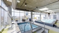 This rooftop-level aquatic amenity space combines industrial design with modern recreational features. The interior is defined by exposed concrete ceilings with visible ductwork and utility conduits, lending an urban, loft-inspired aesthetic. Cylindrical pendant lighting and track-mounted fixtures illuminate the space efficiently while complementing the raw architectural elements overhead.

The facility houses both a hot tub and a swimming pool, finished in blue tile and set into a clean, white-tiled surround. Stainless steel entry rails provide safe access to both water features. The pool area is generously proportioned, offering ample space for lap swimming or leisure.

Floor-to-ceiling windows line the perimeter, flooding the interior with natural light and framing panoramic views of the surrounding urban skyline. The transparent facade seamlessly connects the indoor environment to an adjacent outdoor terrace, which is visible through the glass and equipped with seating and lounge furniture for additional relaxation.

The overall ambiance is contemporary and resort-inspired, balancing the rugged character of exposed structural elements with the polished finish of the aquatic features and the expansive cityscape backdrop.