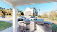 This single-story community clubhouse is designed with an open, welcoming layout highlighted by soaring vaulted ceilings and elegant arched doorways throughout. The interior features light-toned tile flooring that runs seamlessly from the spacious entry foyer into adjoining areas, lending a cohesive and airy feel. Tall decorative planters with lush greenery flank the central archways, enhancing the grand scale of the common areas.

The kitchen is well-appointed with warm wood cabinetry, granite countertops, stainless steel appliances, and an oval island with bar seating — ideal for gatherings and events. Adjacent to the main common area, a dedicated office space offers multiple workstations, wainscoting details, and large windows that overlook outdoor amenities, allowing ample natural light.

Outdoors, a covered patio with white structural columns provides a comfortable shaded retreat, featuring a built-in stone outdoor kitchen equipped with multiple stainless steel grills — perfect for communal entertaining. A secondary covered outdoor area frames tranquil views of mature trees and natural landscaping, offering a serene wooded backdrop. Overall, the space balances functional amenities with an inviting, well-maintained aesthetic suited for community use.