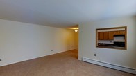 Located at 134 Innis Ave in Poughkeepsie, New York, this single-floor unit offers a straightforward and functional layout designed for comfortable everyday living. The living area is generously proportioned, featuring wall-to-wall carpeting in a neutral tan tone that adds warmth underfoot. Cream-colored walls throughout create a bright, clean aesthetic, while the smooth white ceiling contributes to a sense of openness. A baseboard heating unit runs along the exterior wall, ensuring efficient climate control throughout the colder months. A notable architectural detail is the pass-through window that connects the living area to the kitchen, enhancing the flow between spaces and making the layout feel more open and communicative. The kitchen, visible through the pass-through, features warm wood-toned cabinetry and appears to include a sink, offering a practical and well-organized cooking space. Ceiling-mounted light fixtures provide consistent illumination throughout the unit. The overall ambiance is clean, neutral, and move-in ready, making it a practical and inviting space suitable for a range of living arrangements. The single-floor design ensures easy accessibility and convenience throughout the entire unit.
