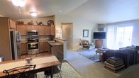 This single-floor residence showcases an open-concept design that seamlessly connects the kitchen and living area, creating a spacious and cohesive living environment. The kitchen is outfitted with warm medium-toned wood cabinetry, dark granite-style countertops, and stainless steel appliances, including a double oven and microwave. A peninsula counter with bar seating provides a natural transition between the kitchen and the adjoining dining area, where a small table and chairs are positioned on hardwood-style flooring. The living area features plush wall-to-wall carpeting and is furnished with a seating arrangement centered around a flat-screen television. Soft, neutral wall colors in warm beige tones carry throughout the space, contributing to a cohesive and inviting ambiance. A window unit air conditioner is visible along one wall, and natural light filters in from windows in the living area. Decorative touches, including framed artwork, table lamps, and accent pieces atop the cabinetry, add warmth and personality to the interior. A hallway visible in the background suggests additional rooms beyond the main living space.