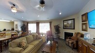 This single-floor space offers a comfortable and well-appointed living area designed with both style and function in mind. The main living room serves as the heart of the space, featuring a classic fireplace with a clean white mantle that anchors the room and provides a natural focal point. Plush, neutral-toned sofas and upholstered armchairs are arranged thoughtfully around a traditional wooden coffee table, creating an inviting seating area ideal for relaxing or entertaining. A ceiling fan with a light fixture is centrally mounted on the textured ceiling, complementing the recessed lighting that adds warmth throughout. Large windows dressed with patterned drapes and horizontal blinds allow generous natural light to fill the room, enhancing the airy feel of the space. Decorative indoor plants add a touch of greenery and life to the corners of the room. Patterned carpeting covers the floor, adding texture and warmth underfoot. A framed wall painting adds a tasteful decorative accent, while a small dining or side table visible near the window suggests a versatile layout. Overall, the space exudes a cozy, traditional ambiance well-suited for comfortable everyday living.