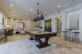 Located in Columbus, GA, this single-floor amenity space combines sophisticated design with versatile functionality. The main gathering area is finished with large-format travertine tile flooring that lends a warm, upscale feel throughout. Abundant natural light filters in through plantation-shuttered windows, complemented by recessed lighting and stylish pendant fixtures overhead.

At the heart of the space sits a billiards table, anchoring the open recreational zone. Adjacent to it is a sleek wet bar featuring a white stone countertop, a richly textured mosaic tile accent wall, and multiple wall-mounted televisions — ideal for social gatherings. Gold-framed bar stools add a contemporary touch. A dedicated lounge area offers plush seating, including a tufted sofa and upholstered armchairs arranged around an area rug, with framed botanical artwork adorning the neutral-toned walls.

A separate media room provides a more intimate experience, outfitted with leather reclining chairs equipped with cup holders, in-wall speakers, and additional framed artwork — creating a private theater-like atmosphere. The space also includes a workstation area with individual desks and chairs for productivity needs. Glass-panel doors and multiple entryways allow for easy flow, while views of the surrounding greenery enhance the overall inviting ambiance.