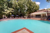 This inviting outdoor pool facility is thoughtfully designed for relaxation and recreation, set within a lush, tree-lined environment that provides both shade and a sense of natural seclusion. The centerpiece is a generously sized rectangular swimming pool with crystal-clear turquoise water, finished with a classic brick coping detail and equipped with stainless steel entry railings for easy access. The pool features a gradual depth beginning at 3½ feet, making it accessible for a range of swimmers.

Surrounding the pool is a spacious concrete sun deck lined with numerous lounge chairs, offering ample space for sunbathing and leisure. A dedicated seating area on a raised patio platform provides additional gathering space, shaded by blue market umbrellas and furnished with bistro-style tables and chairs — ideal for casual outdoor dining or socializing.

The area is fully enclosed by a decorative iron fence with white column posts, providing both safety and an aesthetically pleasing boundary. Ornamental plantings, including flowering crepe myrtles and potted arrangements, add vibrant color throughout the space.

Adjacent to the pool sits a charming single-story clubhouse building clad in neutral siding, featuring a covered front porch with white columns, hanging planters, and welcoming staircase entry — offering a functional and attractive poolside retreat.