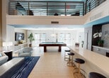 Located at 2263 S Douglas Rd in Miami, FL, this modern amenity lounge delivers a sophisticated social environment designed for both relaxation and entertainment. The expansive open-plan layout features soaring double-height ceilings with an upper-level mezzanine overlooking the main floor, creating a dramatic sense of vertical space. Light wood-look flooring and crisp white walls are accented by bold teal architectural details, lending the space a contemporary coastal energy.

The central entertainment area is anchored by a full-size billiards table and is flanked by a comfortable lounge seating arrangement — including plush sofas and accent chairs in soft aqua tones — set atop a richly patterned navy rug. A large-scale abstract artwork adds a vibrant focal point to this area. Floor-to-ceiling glass doors and large windows flood the space with natural light and provide views of the outdoor surroundings.

The kitchenette is outfitted with dark wood cabinetry, a stainless steel refrigerator, a wall-mounted television, a sink, and a central island with bar seating — ideal for casual gatherings. A dedicated meeting or dining nook features a round wood table with teal upholstered chairs, elegant sheer drapery, and a statement pendant light, offering a more intimate setting within the larger communal space.