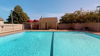 This single-story property showcases a well-appointed outdoor amenity area centered around a large, rectangular swimming pool filled with vivid turquoise water. The pool is notably spacious, offering ample room for both recreational swimming and relaxed lounging. A clearly marked lane line runs along the pool's floor, suggesting suitability for lap swimming as well as leisure use.

Directly behind the pool stands a dedicated pool house structure featuring a pitched, shingled roof and multiple doors, likely providing restroom facilities and storage for pool equipment. The exterior is finished in a neutral tone that complements the surrounding environment. To the left of the pool house, a covered cabana with a distinctive red roof offers a shaded retreat, ideal for escaping the sun during warm days.

The entire pool area is enclosed by a wooden privacy fence, creating a secluded and secure environment. Mature trees line the perimeter, adding natural greenery and additional shade. The surrounding deck provides practical deck space for lounge seating. Under open blue skies, the overall atmosphere of this outdoor space is inviting, private, and well-suited for warm-weather recreation and relaxation.