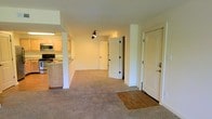 This apartment at Lakewood Park Apartments offers a comfortable and functional single-floor layout designed for everyday living. The open-concept living and dining area is finished with plush wall-to-wall carpeting, creating a warm and inviting atmosphere, while a hardwood-style entryway at the front door provides a clean transition into the home. A track lighting fixture overhead ensures the living space is well-lit throughout. The kitchen is partially separated from the living area by a structural column and a raised counter with a granite-style surface, offering a natural division while maintaining an open feel. The kitchen is equipped with light wood cabinetry, a built-in microwave, and a black range, making it both practical and functional. Interior doors with classic paneling lead to additional rooms and closet spaces, suggesting well-organized storage throughout. The neutral color palette — featuring soft white walls and warm-toned flooring — lends the space a clean, move-in-ready aesthetic. The overall layout is efficient and thoughtfully designed, offering a comfortable living environment within the Lakewood Park Apartments community.