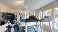Located at 884 Thurber Dr W in Columbus, Ohio, this single-floor space is highlighted by a well-appointed fitness area designed to support an active lifestyle. The exercise room is outfitted with cardio equipment, including a treadmill and an elliptical machine, positioned for easy use and movement. Large glass-paned doors line the room, inviting an abundance of natural light and offering views of the surrounding outdoor area, creating an open and energizing atmosphere. Wall-mounted mirrors expand the sense of space and provide functional support for workouts. A textured ceiling with track lighting ensures the room remains well-lit regardless of the time of day. The light-toned walls and dark flooring provide a clean, functional aesthetic throughout the fitness space. A glimpse through the mirrors reveals an adjacent living area, suggesting a comfortable and versatile layout beyond the workout room. Overall, this space delivers a practical and inviting environment well-suited for health and wellness routines, with thoughtful design details that enhance both functionality and comfort.