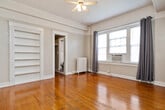This studio is a well-organized single-floor space that makes efficient use of its compact footprint. The living area serves as the heart of the unit, showcasing warm, richly toned hardwood floors that extend throughout. A ceiling fan anchors the room overhead, while a classic cast-iron radiator provides supplemental heating. A projecting bay window fitted with an air conditioning unit allows natural light to fill the space, complemented by gray curtain panels. Built-in open shelving is integrated directly into the wall, offering functional display and storage without consuming floor space. A dedicated closet provides additional storage.

The kitchen is efficiently appointed with white cabinetry, a dark granite-style countertop, a stainless steel sink, and a mosaic tile backsplash featuring a black-and-white geometric pattern. A full-size refrigerator completes the setup.

The bathroom presents a distinct character with bold black-and-white diamond-patterned wall tiles, dark marble-style flooring, a soaking bathtub, and vintage-style exposed plumbing fixtures. A hallway connects the rooms, contributing to a logical and functional flow throughout the space.