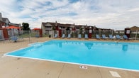 This outdoor amenity space centers around a generously sized swimming pool filled with crystal-clear blue water, creating a refreshing focal point for the surrounding community. The pool area is enclosed by a black iron security fence, providing both safety and a defined boundary for the space. A wide concrete pool deck surrounds the water, offering ample room for movement and relaxation. Numerous lounge chairs are arranged along the poolside, providing residents with comfortable spots to enjoy the open sky and fresh air. A shade umbrella positioned toward one corner adds a practical option for those seeking relief from direct sunlight. Brick pillar accents complement the iron fencing, adding a touch of architectural character to the enclosure. In the background, the multi-unit residential buildings are visible, featuring traditional pitched rooflines, balconies, and brick detailing that contribute to the community's cohesive aesthetic. The surrounding landscape opens up to wide skies, enhancing the sense of openness and outdoor freedom. Overall, the pool area presents a well-organized and inviting recreational space suitable for warm-weather enjoyment.