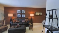 This two-floor home offers a thoughtfully designed interior with a warm and inviting ambiance throughout. The living area makes a strong visual impression with a bold terracotta accent wall that anchors the room and creates a cozy, richly toned atmosphere. Plush wall-to-wall carpeting adds comfort underfoot, while soft ambient lighting from floor lamps enhances the welcoming feel of the space. The living room is furnished with a classic dark leather sofa and matching ottoman, complemented by decorative throw pillows in coordinating earth tones. A curated gallery of framed artwork adorns the accent wall, adding personality and visual interest. Additional framed art on the adjacent wall further contributes to the space's decorated, lived-in character. A staircase leads to the second floor, suggesting additional living or sleeping quarters above. The neutral white ceiling and trim provide a clean contrast to the deeper wall tones, balancing the overall color palette. The layout flows naturally, making the space feel open and connected. Throughout the home, the design choices reflect a consistent aesthetic that emphasizes warmth, comfort, and intentional decorating — making it an appealing and functional residence.
