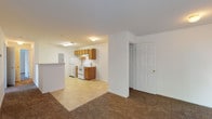 This single-story unit offers a practical and functional layout with clearly defined living spaces. The kitchen is well-equipped with warm-toned oak cabinetry, both upper and lower storage, light-colored countertops, and white appliances including a refrigerator, electric range, and dishwasher. Tile flooring runs throughout the kitchen, which also houses the electrical panel and benefits from overhead fluorescent lighting. The kitchen opens toward the living area, which transitions to plush carpeted flooring and features a sliding glass door that lets in natural light and provides direct access to an outdoor green space. The living area is generously sized and connects to a hallway leading to the rest of the unit. Two full bathrooms are present, each featuring tile flooring, oak vanity cabinetry, white countertops, a large mirror, and a shower-tub combination. A spacious walk-in closet offers wire shelving and carpeted flooring, providing ample storage. Throughout the unit, white walls and neutral finishes create a clean, cohesive aesthetic well-suited to a variety of furnishing styles.