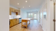 This well-designed single-floor unit offers a clean, contemporary aesthetic throughout. The open-concept kitchen and living area serves as the heart of the space, featuring warm, light wood cabinetry with sleek black hardware, white quartz countertops, and a full suite of stainless steel appliances including a refrigerator, range, microwave, and dishwasher. A freestanding kitchen island with a lower shelf provides additional prep and storage space, seamlessly connecting the kitchen to the living area. Light wood-toned luxury vinyl plank flooring flows throughout the main living spaces, enhancing the bright, airy atmosphere created by recessed lighting and large windows. The living area opens toward a glazed exterior door that allows natural light to pour in and offers views of the surrounding greenery. The bedroom features plush carpeting, a ceiling fan, and double windows with horizontal blinds that frame pleasant outdoor views. A second carpeted room connects directly to the kitchen area and offers flexible use as a bedroom or office. The bathroom is finished with large-format neutral tile in the tub-shower surround, wood-toned vanity cabinetry, and light plank flooring, maintaining the cohesive modern palette found throughout the unit. Overall, the space presents a fresh, functional, and inviting environment with consistent attention to contemporary finishes.