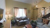 This two-story residence offers a thoughtfully designed interior that balances functionality with a warm, inviting atmosphere. The living room serves as the heart of the main level, characterized by vaulted ceilings that lend a sense of openness and volume to the space. A large window dressed with floral curtains allows generous natural light to fill the room, creating a bright and airy environment. The living area comfortably accommodates seating arranged around a wooden coffee table, complemented by an abundance of indoor plants that add a natural, organic touch to the décor. A pendant light fixture overhead provides ambient illumination, enhancing the room's welcoming character. Adjacent to the living space, a defined entryway features a front door with a decorative rug and framed wall art nearby, establishing a clear transition point into the home. A staircase with a wrought iron railing is visible from the living area, providing access to the second floor. The carpeted flooring throughout the main living space adds warmth and comfort underfoot, while the neutral wall tones create a versatile backdrop suitable for a variety of interior styles.