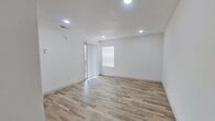 This single-floor, one-bedroom, one-bathroom unit offers an efficient and thoughtfully arranged layout. The combined living and kitchen area forms the heart of the space, featuring light-toned laminate wood flooring that runs throughout. Recessed lighting illuminates the living area, which is brightened further by a sliding glass door and an adjacent window fitted with horizontal blinds, allowing natural light to filter in. The walls throughout are finished in a clean, neutral white, contributing to an open and airy feel.

The galley-style kitchen is positioned adjacent to the living area and is equipped with dark navy cabinetry, white countertops, a stainless steel sink, a dishwasher, and a black electric range. The layout maximizes counter space along both sides of the kitchen corridor, providing practical functionality within a compact footprint.

The bedroom is a well-proportioned, private space featuring plush carpeting, white walls, and a window with blinds that allows natural light in. The bathroom includes a full tub-shower combination with white tile surround, a toilet, and laminate-style flooring, maintaining the consistent, clean aesthetic found throughout the unit. Overall, the space presents a practical and neatly finished interior suited for efficient single-occupant living.