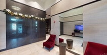 This elegantly designed lobby serves as a striking first impression for residents and visitors alike. The space is defined by soaring ceilings and expansive, light-toned porcelain tile flooring that creates a clean, polished foundation throughout. A dramatic dark marble feature wall commands attention as a bold focal point, its high-gloss surface adorned with a decorative floral arrangement that adds a welcoming, organic touch. Warm beige wall paneling with subtle horizontal banding lines the perimeter, lending the space a refined, contemporary character. Plush crimson velvet accent chairs paired with cylindrical bronze side tables offer an inviting seating area with a rich contrast of color and texture. A built-in digital display screen, set into a niche framed by the stone-toned wall panels, keeps residents informed with building updates. Floor-to-ceiling glass entry doors with sleek black framing allow natural light to filter into the space and provide a seamless visual connection to the exterior. A dedicated mail and package area features traditional mailboxes alongside a modern automated Package Concierge system, set against a stylish plaid-patterned wall covering. Colorful abstract artwork adds a final creative flourish, completing the lobby's upscale and thoughtfully curated ambiance.