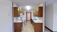 This single-floor space centers around a classic galley-style kitchen designed for practical, efficient use. The kitchen features warm oak cabinetry lining both sides of the corridor-style layout, providing ample upper and lower storage throughout. The cabinetry is finished with dark hardware accents, complementing the wood tones and adding a consistent visual detail across the space.

White laminate countertops run the length of both sides, offering generous workspace. On the left side, a white electric range with a built-in overhead microwave serves as the primary cooking station. On the right side, a double basin sink sits beneath additional upper cabinetry, accompanied by a standard faucet fixture. A white refrigerator is positioned at the far end of the left wall, making efficient use of the available space.

The flooring consists of neutral, light-toned vinyl tile, which is easy to maintain and complements the overall palette of the kitchen. A textured backsplash lines the walls between the countertops and upper cabinets on both sides, adding subtle depth. Overhead, a flush-mount light fixture provides consistent illumination. A doorway at the far end of the kitchen leads into an adjacent room, contributing to the overall flow of the floor plan.