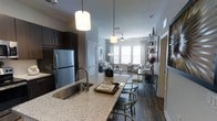 This single-floor residence showcases a thoughtfully designed open-concept layout that seamlessly connects the kitchen, dining, and living areas. The kitchen serves as a focal point, featuring granite or stone-composite countertops, dark espresso cabinetry, stainless steel appliances including a full-size refrigerator, and a large island with an undermount sink and pendant lighting overhead. Bar-style seating along the island creates a natural transition into the dining space.

The living area is bathed in natural light through a bank of large windows dressed with white blinds, creating a bright and airy atmosphere. A ceiling fan is centrally positioned in the living room, enhancing both comfort and functionality. Rich hardwood-style flooring runs throughout the space, complementing the neutral grey wall tones and modern décor.

An open shelving unit provides decorative display space near the living area, while a louvered door adds a classic architectural detail. The overall ambiance is contemporary and refined, combining dark and light contrasts for a balanced, inviting interior.