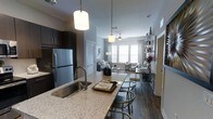 This single-floor residence showcases a thoughtfully designed open-concept layout that seamlessly connects the kitchen, dining, and living areas. The kitchen serves as a focal point, featuring granite or stone-composite countertops, dark espresso cabinetry, stainless steel appliances including a full-size refrigerator, and a large island with an undermount sink and pendant lighting overhead. Bar-style seating along the island creates a natural transition into the dining space.

The living area is bathed in natural light through a bank of large windows dressed with white blinds, creating a bright and airy atmosphere. A ceiling fan is centrally positioned in the living room, enhancing both comfort and functionality. Rich hardwood-style flooring runs throughout the space, complementing the neutral grey wall tones and modern décor.

An open shelving unit provides decorative display space near the living area, while a louvered door adds a classic architectural detail. The overall ambiance is contemporary and refined, combining dark and light contrasts for a balanced, inviting interior.