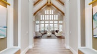 Located in Port Saint Lucie, FL, this elegantly appointed single-floor space offers a generous and well-connected layout encompassing a living room, dining room, and kitchen. The architectural centerpiece is the dramatic vaulted ceiling with exposed wood beams and a striking gold chandelier, creating an airy, grand atmosphere that immediately captures attention upon entry. Walls of floor-to-ceiling windows flood the space with natural light while providing open views of the surrounding outdoor areas, including a pool and landscaped grounds. Light-toned wood-look tile flooring flows seamlessly throughout, unifying the space with a warm, contemporary aesthetic. The living area is anchored by a sleek wood-paneled feature wall hosting a mounted flatscreen TV above a modern linear fireplace, flanked by gold sconce lighting. Comfortable lounge seating is thoughtfully arranged to encourage conversation and relaxation. The kitchen is a standout, showcasing crisp white cabinetry, a large quartz-topped island with bar seating, bold gold cylinder pendant lights, and a textured tile backsplash. A charming built-in dining nook with a cobalt blue accent wall and art lighting adds a distinctive, boutique touch. The dining area, bathed in natural light, completes this inviting and functionally rich space.