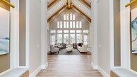 Located in Port Saint Lucie, FL, this elegantly appointed single-floor space offers a generous and well-connected layout encompassing a living room, dining room, and kitchen. The architectural centerpiece is the dramatic vaulted ceiling with exposed wood beams and a striking gold chandelier, creating an airy, grand atmosphere that immediately captures attention upon entry. Walls of floor-to-ceiling windows flood the space with natural light while providing open views of the surrounding outdoor areas, including a pool and landscaped grounds. Light-toned wood-look tile flooring flows seamlessly throughout, unifying the space with a warm, contemporary aesthetic. The living area is anchored by a sleek wood-paneled feature wall hosting a mounted flatscreen TV above a modern linear fireplace, flanked by gold sconce lighting. Comfortable lounge seating is thoughtfully arranged to encourage conversation and relaxation. The kitchen is a standout, showcasing crisp white cabinetry, a large quartz-topped island with bar seating, bold gold cylinder pendant lights, and a textured tile backsplash. A charming built-in dining nook with a cobalt blue accent wall and art lighting adds a distinctive, boutique touch. The dining area, bathed in natural light, completes this inviting and functionally rich space.