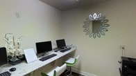 This single-floor space functions as a dedicated business center, thoughtfully designed to offer a focused and functional environment for work and productivity. The room features a sleek, granite-topped countertop desk that runs along the wall, accommodating two desktop computer workstations complete with monitors, keyboards, and mice — providing convenient access for users needing to handle business tasks, correspondence, or browsing. Modern green-and-white adjustable bar-style chairs are positioned at the workstations, adding a contemporary pop of color against the neutral, light-toned walls. A decorative sunburst-style mirror with a metallic finish serves as a stylish focal point on the wall, elevating the overall aesthetic of the space. The room features a textured ceiling and dark baseboards that provide clean contrast to the light walls. The compact layout is efficiently utilized, making the most of the available space while maintaining an uncluttered, welcoming atmosphere. Overall, the business center strikes a balance between practicality and design, offering a polished setting suited for professional tasks in a shared or hospitality-oriented environment.
