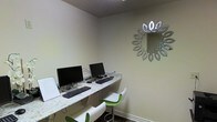 This single-floor space functions as a dedicated business center, thoughtfully designed to offer a focused and functional environment for work and productivity. The room features a sleek, granite-topped countertop desk that runs along the wall, accommodating two desktop computer workstations complete with monitors, keyboards, and mice — providing convenient access for users needing to handle business tasks, correspondence, or browsing. Modern green-and-white adjustable bar-style chairs are positioned at the workstations, adding a contemporary pop of color against the neutral, light-toned walls. A decorative sunburst-style mirror with a metallic finish serves as a stylish focal point on the wall, elevating the overall aesthetic of the space. The room features a textured ceiling and dark baseboards that provide clean contrast to the light walls. The compact layout is efficiently utilized, making the most of the available space while maintaining an uncluttered, welcoming atmosphere. Overall, the business center strikes a balance between practicality and design, offering a polished setting suited for professional tasks in a shared or hospitality-oriented environment.