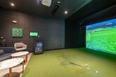 This indoor golf simulator space is a compact, single-floor venue designed to deliver an immersive golfing experience. The simulator bay is the centerpiece, featuring a large high-definition projection screen that displays realistic course visuals, complemented by green turf flooring that extends from the hitting area to create an authentic feel underfoot. The walls of the bay are finished in dark tones, minimizing distractions and enhancing the visual impact of the projected course.

Adjacent to the simulator bay is a well-appointed lounge area that blends functionality with refined aesthetics. Dark, textured wallcovering with a subtle pattern runs throughout, establishing a sophisticated and moody ambiance. The lounge is furnished with a cushioned armchair, a modern swivel chair, and a round marble-top side table, providing a comfortable setting for players and guests between sessions. A wall-mounted monitor allows viewers to follow gameplay from the seating area, while a dedicated simulator control station with a touchscreen interface sits nearby for easy course selection and settings management. Golf bags and clubs are stored conveniently within the space. Large glass panels on one side of the room allow natural light to filter in from an adjoining area, offering a visual connection to the broader facility while maintaining the intimate, focused atmosphere of the simulator suite.