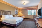 This well-appointed studio suite in Rothschild, Wisconsin, offers a thoughtfully designed single-floor layout that combines living, sleeping, dining, and cooking areas into one cohesive space. The interior embraces a warm lodge aesthetic, highlighted by natural wood accents including pine crown molding, a distinctive twig-crafted headboard, and rustic hickory-style dining chairs and décor. Warm earth tones — soft yellows and olive greens — complement the light wood flooring and patterned carpeting throughout the main area.

The sleeping area is anchored by a king-sized bed dressed in crisp white linens, with wall-mounted sconce lighting and a wooden nightstand for convenience. Adjacent to the bed, a full kitchenette is tucked into a recessed nook, equipped with dark wood cabinetry, a stainless steel refrigerator, microwave, stove, dishwasher, and a sink with dark countertops — ideal for extended stays. A dining table with rustic twig-back chairs seats up to four and sits beside a flat-screen TV on a wooden media dresser, completing the living area.

Natural light filters in through a window at the far end of the room, adding brightness to the inviting atmosphere. The bathroom features light hardwood-style flooring and a walk-in shower with elegant marble-style tile walls. The entrance area provides a smooth transition into this comfortable and characterful space.