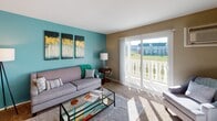 This single-floor residence offers a well-organized layout that balances comfort and functionality throughout its key living areas. The living room serves as the centerpiece of the home, featuring plush carpeting, a bold teal accent wall adorned with decorative artwork, and large sliding glass doors that open onto a private balcony. The balcony provides pleasant outdoor views of the surrounding green landscape and neighboring buildings, allowing natural light to flood the space throughout the day. The bedroom is a standout room, distinguished by a striking burnt-orange accent wall that adds warmth and energy to the space. A set of mirrored sliding closet doors enhances the sense of depth and provides generous storage. A window allows natural light to brighten the room, creating an inviting and restful atmosphere. The bathroom is neatly appointed with warm-toned ceramic tile flooring, wood-cabinet vanity storage, a single-sink countertop, and a full bathtub with a shower combination. The kitchen features warm oak cabinetry, a double stainless steel sink, light-toned countertops, and tile flooring. A full-size refrigerator completes the functional cooking space. Overall, the unit presents a cohesive and colorful interior design with practical amenities throughout.