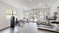 Located at 450 Technology Pkwy NW in Norcross, Georgia, this fitness center at Spoke at Peachtree Corners is a thoughtfully designed, single-floor facility built to support a comprehensive workout experience. The space features crisp white walls, crown molding, and recessed lighting throughout, creating a clean, bright, and motivating environment. Durable speckled rubber flooring provides comfort and safety across the entire workout area.

The cardio zone is well-appointed with treadmills, an elliptical trainer, and a stationary spin bike, all equipped with digital displays for performance tracking. A dedicated strength training area houses a multi-function cable machine, a leg extension/curl machine, a dumbbell rack with a full range of weights, resistance bands, and exercise mats mounted to the wall. A sled training platform adds a functional fitness element to the space.

Large windows allow ample natural light to fill the room, offering views of the surrounding greenery and outdoor amenities. Glass-paneled double doors provide direct access to the outdoor area, seamlessly connecting the interior fitness space to the exterior environment. A glass partition wall offers a view into an adjacent lounge or common area, enhancing the open, connected feel of the facility overall.