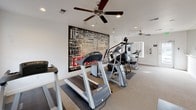 This single-level fitness center is designed to support a well-rounded workout experience in a clean, bright, and functional environment. The space is divided into two distinct exercise zones, each outfitted with ceiling fans to promote air circulation during workouts.

The cardio area houses multiple Matrix treadmills and an elliptical machine, positioned near large windows that allow natural light to fill the room. A glazed entry door opens to the exterior, offering views of the surrounding grounds. Safety exit signage and security features are also present throughout.

Adjacent to the cardio zone, a dedicated strength and conditioning area features a StairMaster climber, a stationary exercise bike, and a multi-function cable resistance machine. This section is anchored by a bold, large-scale typographic mural covering an entire wall, displaying fitness-themed words such as "Endurance," "Intensity," "Cardio," and "Strength," adding a motivating visual energy to the space.

A separate stretch and movement area lines one wall with full-length mirrors and a ballet-style barre, ideal for stretching, yoga, or low-impact exercise. This reflective wall visually expands the room and enhances the open, airy feel of the space. Recessed lighting and neutral-toned carpet complete the clean, purpose-driven aesthetic throughout.