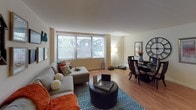This single-floor residence offers a thoughtfully arranged layout with distinct living, dining, sleeping, and cooking areas. The living room serves as the central gathering space, anchored by a large window fitted with vertical blinds that invite abundant natural light and offer outdoor views. Warm hardwood-style flooring runs throughout, lending continuity and a welcoming feel to the space.

The living area flows naturally into a dedicated dining space, creating an open, connected atmosphere well-suited for everyday living and casual entertaining. A galley-style kitchen lines one wall with rich wood cabinetry, dark granite-look countertops, a mosaic tile backsplash, and black appliances including a full-sized refrigerator and dishwasher. The efficient layout maximizes storage and workspace.

The bedroom is generously proportioned, featuring a large window that continues the home's bright aesthetic. A built-in closet with sliding doors provides organized storage, while the hardwood-style flooring carries through from the main living area. The room comfortably accommodates a full bedroom suite.

The bathroom is finished with large-format gray tiles, a glass-enclosed shower-tub combination with a decorative mosaic accent border, a pedestal sink, and warm overhead lighting. Mirrored storage and built-in shelving add practical functionality to the refined design.