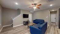 This two-floor student unit in Memphis, Tennessee is designed with functionality and comfort in mind, making it well-suited for student living. Throughout the space, light-toned luxury vinyl plank flooring provides a cohesive, low-maintenance foundation, complemented by neutral taupe-toned walls and consistent ceiling fans with integrated lighting in each room.

The main living area on the ground floor features a staircase, a wall-mounted flat-screen television, and seating arranged around a central coffee table. Adjacent to the living space is a dedicated dining area with a table and chairs, benefiting from a window that allows natural light. The kitchen is a notable feature, offering dark espresso cabinetry, stainless steel appliances including a microwave and range, white countertops, and a peninsula with bar seating for four. A window above the sink brings in additional natural light.

A dedicated laundry room houses a full-size washer and dryer alongside upper cabinetry and counter space. The bathroom includes a dark wood vanity with a white countertop and a well-lit mirror. The bedroom accommodates a bed frame and desk area, with a window providing natural light and a wall-mounted mini-split HVAC unit ensuring individual climate control.