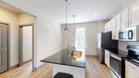 This single-floor, one-bedroom, one-bathroom residence is designed with a clean, contemporary aesthetic and a practical, well-organized layout. The kitchen is a standout feature, showcasing dark granite countertops, a central island with a stainless steel undermount sink and a sleek pull-down faucet, and crisp white shaker-style cabinetry extending to the ceiling for ample storage. A full suite of stainless steel appliances — including a refrigerator, electric range, built-in microwave, and dishwasher — adds a polished, modern touch. Stylish industrial-style pendant lights hang above the island, complementing the light wood-look flooring that flows throughout the main living areas. The living space is bright and airy, featuring large windows with blinds that invite natural light, a ceiling fan, and neutral-toned walls that create a calm, versatile backdrop. The bedroom continues the neutral palette with plush carpeting, a ceiling fan, and a window offering natural light. The bathroom is finished with white subway tile surrounding a soaking tub and shower combo, wood-look tile flooring, a vanity with a dark countertop, and a window that brightens the space naturally.