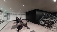 This fitness center is designed with a contemporary aesthetic, combining functionality with bold visual branding across its single-floor layout. The space is divided into distinct workout zones, each thoughtfully equipped to support a comprehensive training experience.

The cardio area features a row of Matrix treadmills and elliptical trainers positioned against a dramatic matte-black accent wall, with recessed and linear LED lighting illuminating the light-toned flooring. A full-length mirror amplifies the sense of space and reflects motivational typography displayed across the walls. Adjacent to this zone, a dedicated cycling area houses multiple Peloton spin bikes alongside a standing elliptical, set against the same striking black wall bearing the phrase "Stronger Than Yesterday."

The strength training zone offers a well-organized selection of selectorized machines, free weight benches, and a rack of dumbbells, all arranged on warm wood-grain flooring beneath wall-mounted televisions and softly lit windows with frosted blue panels.

A glass-enclosed studio serves as a separate functional training space, its walls decorated with bold typographic graphics referencing cardio, stretching, weight lifting, and nutrition. The transparent glass partitions maintain visual connectivity throughout the facility while defining individual training environments, contributing to an open, motivating atmosphere.