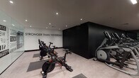 This fitness center is designed with a contemporary aesthetic, combining functionality with bold visual branding across its single-floor layout. The space is divided into distinct workout zones, each thoughtfully equipped to support a comprehensive training experience.

The cardio area features a row of Matrix treadmills and elliptical trainers positioned against a dramatic matte-black accent wall, with recessed and linear LED lighting illuminating the light-toned flooring. A full-length mirror amplifies the sense of space and reflects motivational typography displayed across the walls. Adjacent to this zone, a dedicated cycling area houses multiple Peloton spin bikes alongside a standing elliptical, set against the same striking black wall bearing the phrase "Stronger Than Yesterday."

The strength training zone offers a well-organized selection of selectorized machines, free weight benches, and a rack of dumbbells, all arranged on warm wood-grain flooring beneath wall-mounted televisions and softly lit windows with frosted blue panels.

A glass-enclosed studio serves as a separate functional training space, its walls decorated with bold typographic graphics referencing cardio, stretching, weight lifting, and nutrition. The transparent glass partitions maintain visual connectivity throughout the facility while defining individual training environments, contributing to an open, motivating atmosphere.