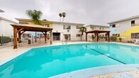 Located at 10237 Western Ave in Downey, California, this outdoor communal pool area is a well-appointed recreational space designed for relaxation and leisure. The centerpiece is a generously sized swimming pool with vivid turquoise water, accessible via a stainless steel entry railing and surrounded by a wide concrete pool deck that offers ample space for lounging and socializing.

Two wooden gazebos with pitched metal roofs anchor opposite ends of the pool deck, each providing shaded seating areas furnished with white benches and chairs. Additional seating is distributed throughout the patio area, including a white outdoor table-and-chair set, inviting casual gatherings and outdoor dining.

The pool area is enclosed by a metal fence, providing a defined and secure boundary. Lush tropical landscaping, including tall palm trees and other mature plantings, frames the space and contributes to a distinctly Southern California atmosphere. The surrounding two-story building, with its white stucco exterior and accents of yellow and pink, forms a courtyard-like backdrop that adds character to the setting.

Overall, the space conveys a relaxed, resort-inspired ambiance well-suited to the warm Southern California climate, offering residents a comfortable and attractive outdoor retreat.