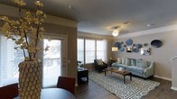 This two-story space in Plano offers a well-designed living area that balances comfort and style. The main living space features hardwood-style flooring and neutral-toned walls accented by crown molding, giving the room a polished, finished look. A ceiling fan with a light fixture provides both illumination and airflow, complementing the abundant natural light that filters in through multiple large windows fitted with horizontal blinds. The windows offer views of an outdoor area, and a glass-panel door provides direct access to what appears to be a private balcony or patio, further enhancing the connection to the outdoors. The living room is generously proportioned, comfortably accommodating a sectional sofa, accent chairs, and a coffee table centered on a bold geometric area rug. Decorative wall art adds a contemporary touch to the space. The open layout encourages a seamless flow between seating and dining areas, making it well-suited for both everyday living and entertaining. With two floors, the space offers versatile living arrangements and additional room beyond what is visible in the main living area.
