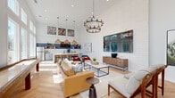 Located at 555 Linger Longer Dr on Johns Island, SC, this single-floor community space delivers an airy, open-plan layout designed for both relaxation and socializing. Soaring ceilings amplify the sense of openness throughout, while floor-to-ceiling windows along one full wall flood the interior with abundant natural light and frame lush outdoor views of the surrounding landscape. The herringbone-patterned light hardwood flooring adds a refined architectural detail that flows seamlessly across the entire space. The lounge area is anchored by a tan leather sectional sofa, modern accent chairs, and a sleek coffee table, all centered beneath a striking two-tiered wrought-iron chandelier. A large wall-mounted display and shiplap feature wall serve as a sophisticated focal point. A shuffleboard table positioned near the windowed wall adds a recreational element to the space. The kitchenette is well-appointed with white subway tile backsplash, navy accent tile, floating wood shelves, stainless steel appliances, quartz countertops, and a dedicated coffee station. Curated artwork displayed at height adds personality and visual interest. The space opens to an adjacent outdoor patio area, enhancing its versatility as a vibrant, community-centered gathering destination.