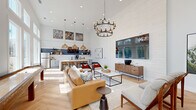 Located at 555 Linger Longer Dr on Johns Island, SC, this single-floor community space delivers an airy, open-plan layout designed for both relaxation and socializing. Soaring ceilings amplify the sense of openness throughout, while floor-to-ceiling windows along one full wall flood the interior with abundant natural light and frame lush outdoor views of the surrounding landscape. The herringbone-patterned light hardwood flooring adds a refined architectural detail that flows seamlessly across the entire space. The lounge area is anchored by a tan leather sectional sofa, modern accent chairs, and a sleek coffee table, all centered beneath a striking two-tiered wrought-iron chandelier. A large wall-mounted display and shiplap feature wall serve as a sophisticated focal point. A shuffleboard table positioned near the windowed wall adds a recreational element to the space. The kitchenette is well-appointed with white subway tile backsplash, navy accent tile, floating wood shelves, stainless steel appliances, quartz countertops, and a dedicated coffee station. Curated artwork displayed at height adds personality and visual interest. The space opens to an adjacent outdoor patio area, enhancing its versatility as a vibrant, community-centered gathering destination.