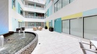 The Resident Atrium is a thoughtfully designed enclosed courtyard that serves as a central communal hub within the building. The space is framed by multi-story facades adorned with a bold, cheerful palette of teal, sky blue, and soft yellow paneling, creating an energetic yet welcoming atmosphere. Large floor-to-ceiling windows and sliding glass doors line the surrounding walls, allowing abundant natural light to filter throughout while providing visual connectivity between interior units and the atrium below.

At the heart of the atrium stands a circular mosaic-tiled fountain, featuring multiple water jets and an illuminated water wall backdrop that adds a dynamic, calming focal point to the space. The light-toned large-format tile flooring extends throughout, lending the area a clean, polished appearance and a sense of openness.

Multiple seating arrangements are distributed across the atrium, including teak-framed lounge chairs with plush white cushions gathered around a central coffee table atop a light blue area rug, as well as modern dining-style chairs paired with a sleek white table for more structured use. Dark rectangular planters with neatly trimmed greenery are positioned throughout, introducing natural elements. Access to the atrium is provided through a teal-accented glass entry door, clearly marked for resident use.